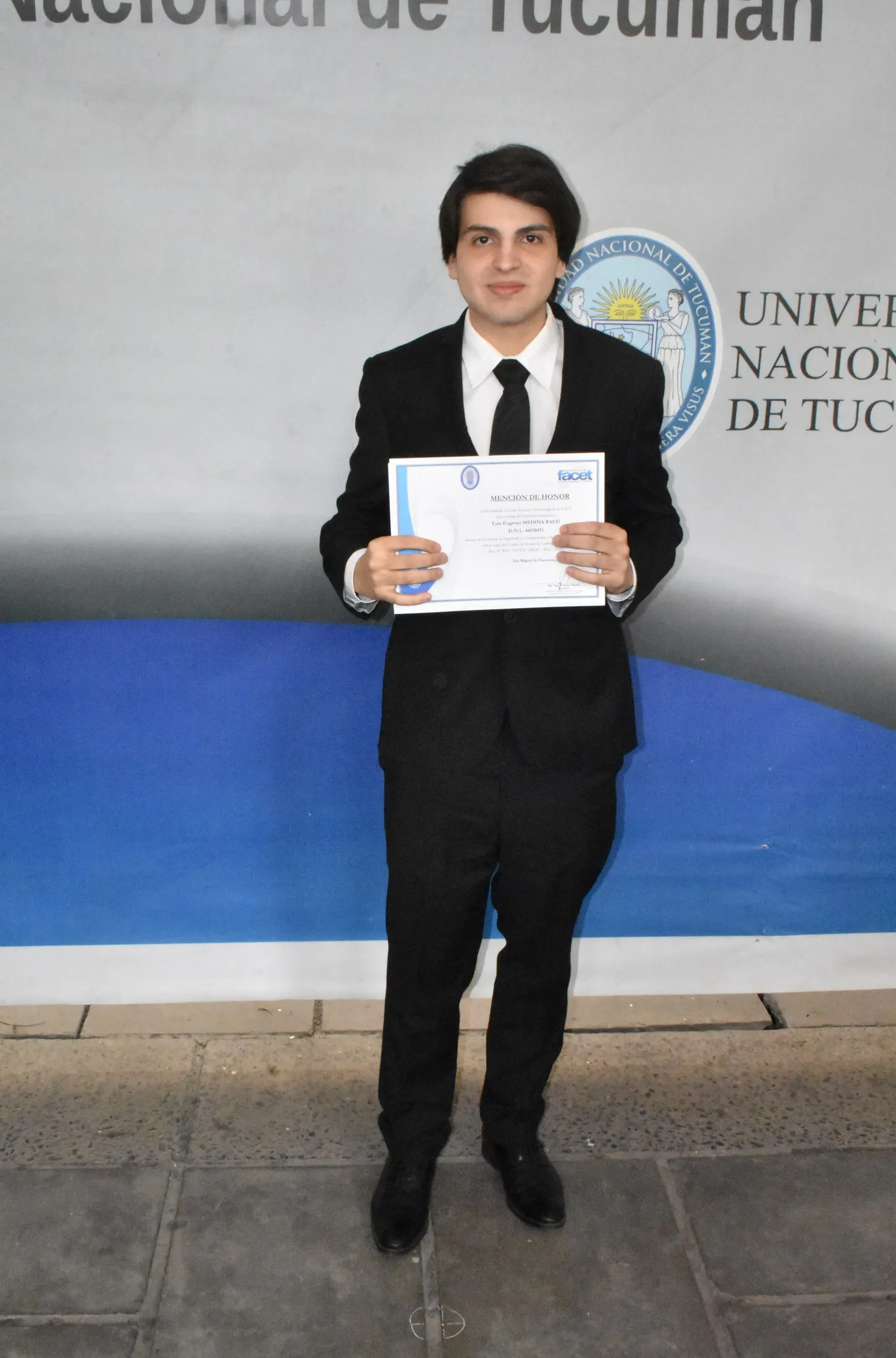 Luis Medina Raed, en pied, tenant la Mention d'honneur devant la banderole de l'Université Nationale de Tucumán (FACET).