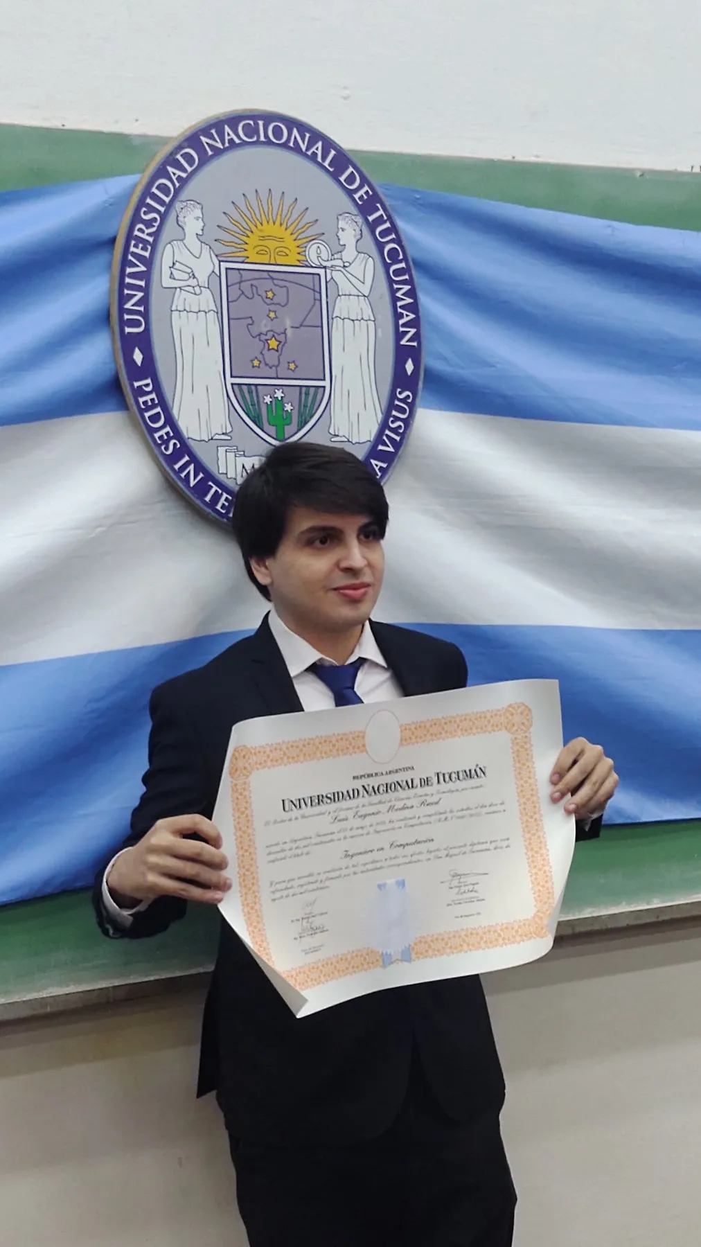 Luis Medina Raed sosteniendo su título frente al escudo de la Universidad Nacional de Tucumán.